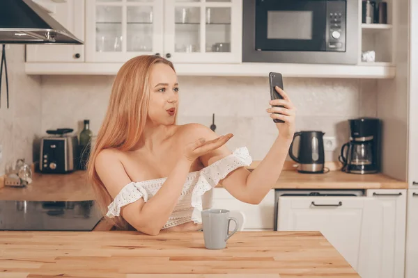 Beautiful woman sits at a table on the background of the kitchen at home. The girl looks at the camera of the phone, keeps her blog, shoots a video, speaks by video communication, smiles. Sends a kiss
