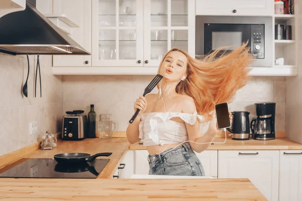 Woman singing and dancing in the kitchen with a smartphone, music.A girl in headphones with a phone stands in the kitchen and prepares food behind an induction stove.