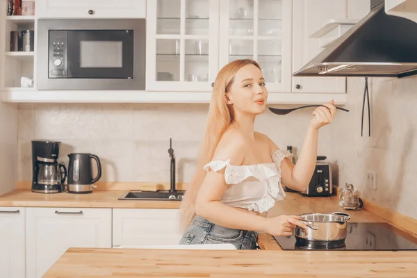 Güzel kız indüksiyon sobasında yemek hazırlıyor. Bir kadın tencerede çorba ya da sos pişirir. Parlak mutfaktaki sarışının.