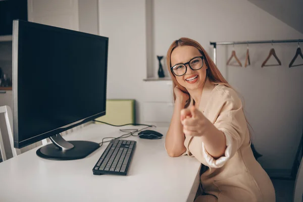 Works behind the monitor. Woman working from home, freelancing, teaching student. A beautiful blonde in glasses and a jacket looks at the camera and points her finger at you. Online training