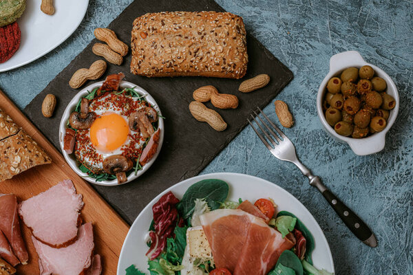 Много еды и блюд на заднем плане. Мясное блюдо и сыр Камамбер с багетом. Салат с прошутто. Оливки. Обжаренные яйца заправить грибами и специями. Веганские овощные котлеты и арахис. Вилка. Доставка продуктов питания.