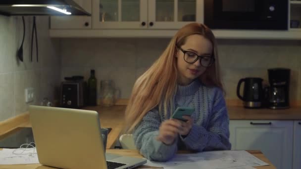 一个女人在家里用笔记本电脑工作 一个穿着毛衣和眼镜的漂亮的金发姑娘 桌上有一个杯子和一张纸 在家上学 上网工作 一个戴耳机的学生通过视频通话 — 图库视频影像