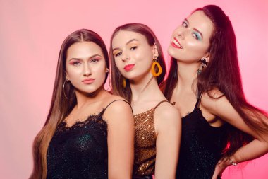 Pembe arka planda gülüp poz veren 3 kız. Farklı renkte makyajlı, parlak elbiseli kızlar. Üç esmer kız dans edip hayatın tadını çıkarıyor. 8 Mart, Kadın Gücü, Kadınlar Günü