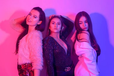 Üç güzel kız neon ışıkta dans ediyor. Parlak elbiseli kızlarla inanılmaz eğlenceli bir parti, bayram kadınlarının gününü kutlamak, 8 Mart, gülümsemek, gülmek, dans etmek, şarkı söylemek. Gece kulübü..