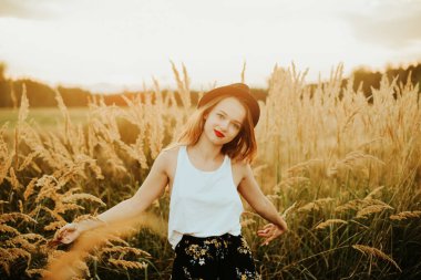 Günbatımı arka planında, buğday tarlasında muhteşem bir kadın. Uzun saçlı şık bir kız neşe saçar, güler, hayattan ve yazdan zevk alır, doğa, mutluluk. Ormanda şapkalı bir manken..