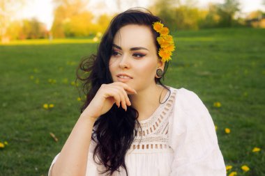 Muhteşem esmer, çimenlerin üzerinde beyaz bir elbiseyle oturuyor ve elinde karahindibalar, sarı karahindiba çiçekleri var. Parlak, şık, sarı makyajlı bir kızın portresi. Kızın artık alerjisi yok..