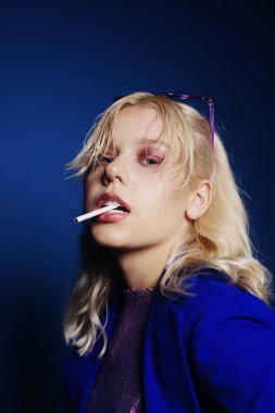 70 'ler, 80' ler, 90 'lar tarzında bir kadın. Parlak gözlüklü ve sigaralı sarışın bir kadın sandalyede oturuyor. Kadın SSCB tarzı. Parlak mavi bir ceket, mor gözlükler, bir kadın sigara içiyor. Grunge tarzı.