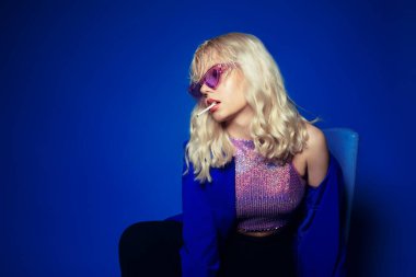 70 'ler, 80' ler, 90 'lar tarzında bir kadın. Parlak gözlüklü ve sigaralı sarışın bir kadın sandalyeye oturmuş, bacaklarıyla. Parlak mavi bir ceket, mor gözlükler, bir kadın sigara içiyor. Grunge tarzı. Mavi arkaplanda moda fotoğrafı