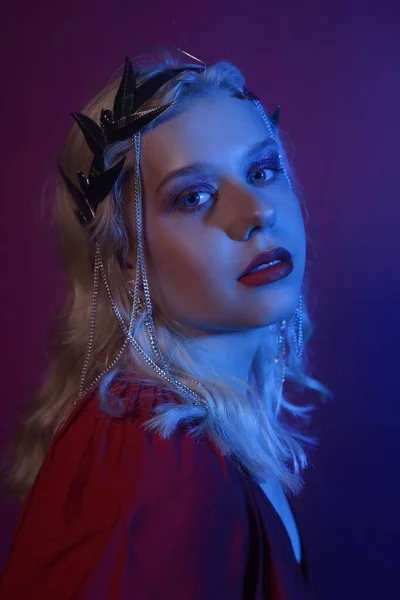 Portrait in neon of a woman in a silver crown with chains. A girl as a princess, queen, princess. Fashion shooting a portrait in red and blue neon. The model is beautiful.