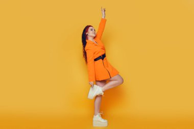 Sarı arka planda güzel bir kız. Turuncu elbiseli ve uzun saçlı bir esmer poz veriyor, dans ediyor, gülüyor, eğleniyor. Stüdyoda sıcak yaz fotoğrafı. Neşe ve duygu