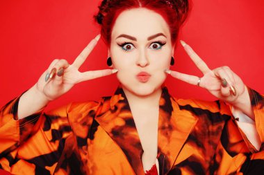 Kırmızı arka planda güzel bir kız. Kızıl saçlar ve kulaklarda tüneller, kırmızı elbise ve dudaklar. Anime poz veriyor. Göz kalemi ve kafasında iki çörek olan kız suratını buruşturur, gülümser, eğlenir. Barış hareketi