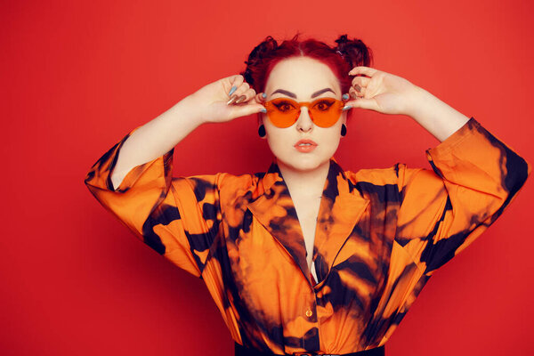 Cute girl on a red background. Red hair and tunnels in the ears, red dress and lips. Anime poses. A girl in orange sunglasses and two buns on her head looks at the camera.