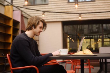 Terasta bir restoranda kahve içen bir adam. Dışarıda dizüstü bilgisayarı olan serbest çalışan, kruvasanlarla kahvaltı yapan biri. İş adamı görünüşü ve hayalleri. Dizüstü bilgisayarlı adam