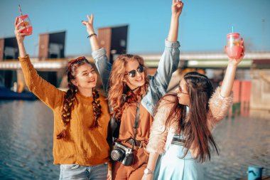 Nehir kıyısında gökyüzüne karşı eğlenen üç kız. Güneş gözlüklü moda kızları. Zıpla, sevin, içki iç, gülümse, eğlenmene bak. Renkli giyinen arkadaşlar yaz havası geçirirler, dans ederler.