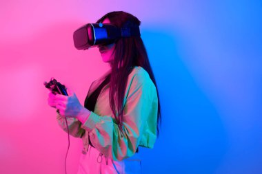 Gülen, neonlu, VR gözlüklü sürpriz kız elinde bir oyun platformu tutuyor ve oyunlar oynuyor. Sanal dünya, teknoloji.