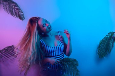 Dans partisi yaz havası. Neon pozlar veren bir model. Mayo giymiş bir kızın moda fotoğrafı ve pembe-mavi ışık altında şortu. Tropikal yaprakların arka planında bir kız yaz fotoğrafı. 