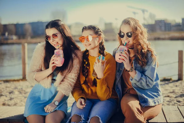 Three girls sit on the beach , near the river, talk, gossip, joke, laugh, smile at womans day. Girls on a hot day eat ice cream. Beauty model wearing sunglasses at summer day. Girl with hairslyle