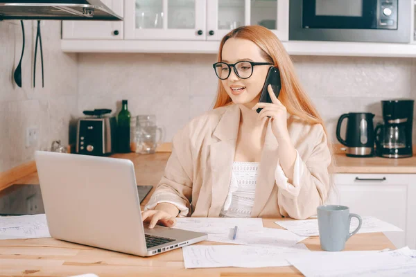 The woman at the laptop works from home in the call center. A beautiful blonde in a jacket and glasses looks into a laptop and writes something down. Girl speaks on the phone and processes the order.