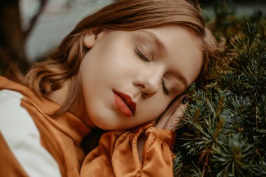 Ahşap, doğa, yeşillik ve ladin arka planında güzel bir model. Kısa saçlı ve kahverengi elbiseli bir kız. Sarışın bir ağaçta yatıyor, manken şık bir şekilde poz veriyor..