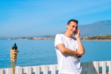 Telefonda konuşan bir adam sahilin, okyanusun ve denizin arka planında konuşuyor. Tatilde olan beyaz gömlekli bir adam. Serbest çalışan arar, gülümser, gezer ve sohbet eder. Seyahat.
