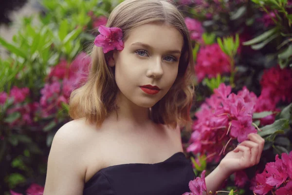 長いピンクのドレスの女の子がピンクの花を背景に芝生の上に庭に座っています 短髪のブロンド ファッショナブルなプリンセス 彼女の髪の花 カメラを見て — ストック写真