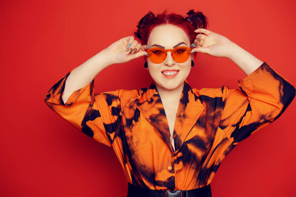 Cute girl on a red background. Red hair and tunnels in the ears, red dress and lips. Anime poses. A girl in bright sunglasses and two buns on her head grimaces, smiles, has fun.