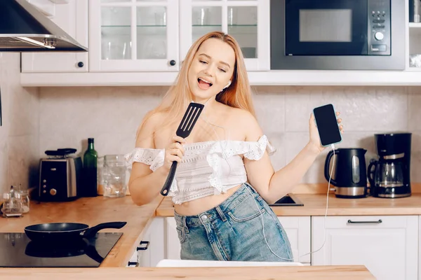 Woman singing and dancing in the kitchen with a smartphone, music.A girl in headphones with a phone stands in the kitchen and prepares food behind an induction stove