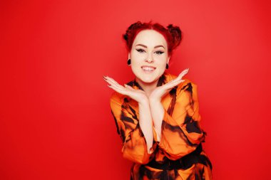 Kırmızı arka planda sevimli bir kız. Kızıl saçlar ve kulaklarda tüneller, kırmızı elbise ve dudaklar. Anime poz veriyor. Göz kalemi ve kafasında iki çörek olan kız suratını buruşturur, gülümser, eğlenir. Japon kültürü