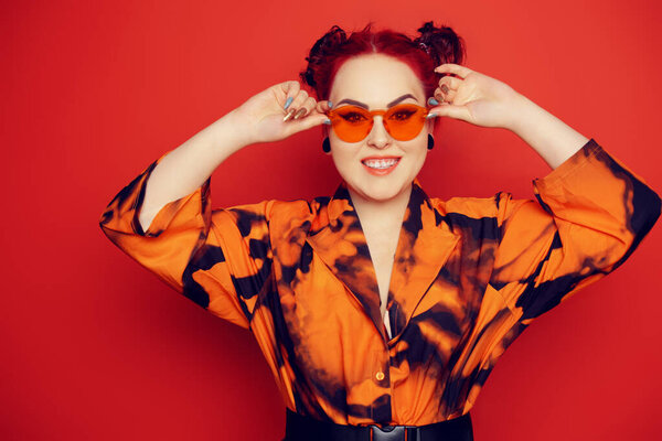 Cute girl on a red background. Red hair and tunnels in the ears, red dress and lips. Anime poses. A girl in bright sunglasses and two buns on her head grimaces, smiles, has fun.