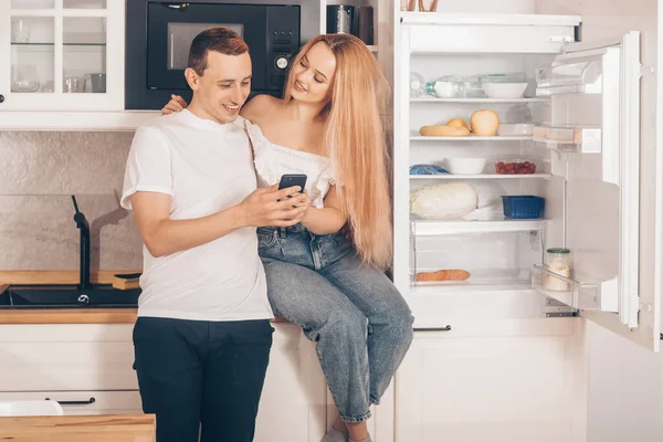 Ordering food through the online store. A couple of lovers in the kitchen at home near the refrigerator. Man and Woman are shopping online through the phone. Smart refrigerator.