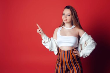 Stüdyoda kırmızı arka plandaki güzel kız boş alanın yan tarafını gösteriyor. Modaya uygun bir kadın gülümser ve kameraya bakar, muhteşem bir gülümseme, mutluluk, neşe.