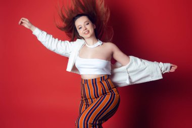 Stüdyoda kırmızı arka planda şık şeyler içinde dans eden güzel enerjik bir kız. Eğlence, mutluluk, dans, hareket, eğlence. Kadın gülümser ve kameraya bakar, yaz fotoğrafı. Uzun saçlı.