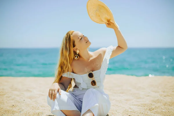 The girl closes herself from the sun and heat with a hat. Fashionable earrings and a beautiful white outfit. The blonde is influenced by the weather on the beach. Girl sunbathing on the sand