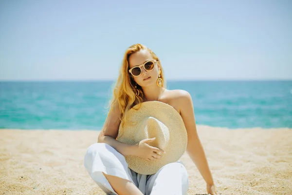 海や太陽を背景にビーチで女の子のファッション写真は 女の子は帽子で彼女の体をカバーしています 大きなイヤリングと白いスーツを着た美しい女性 — ストック写真
