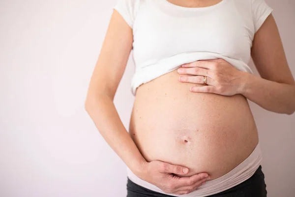 Pregnant woman wearing a white t-shirt with hands on belly on blank background. Central view. Background with copy space.