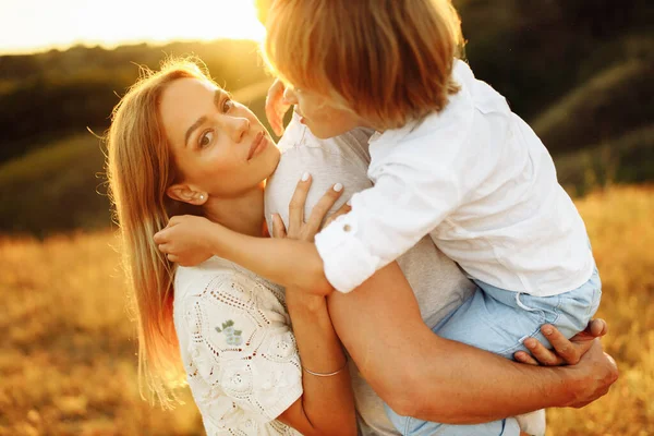 Genitori adorabili con figlio gioioso che cammina sul campo, padre premuroso che tiene il figlio prescolare sulle braccia, madre sorridente e amorevole che abbraccia delicatamente padre bello, genitorialità e concetto di infanzia — Foto Stock