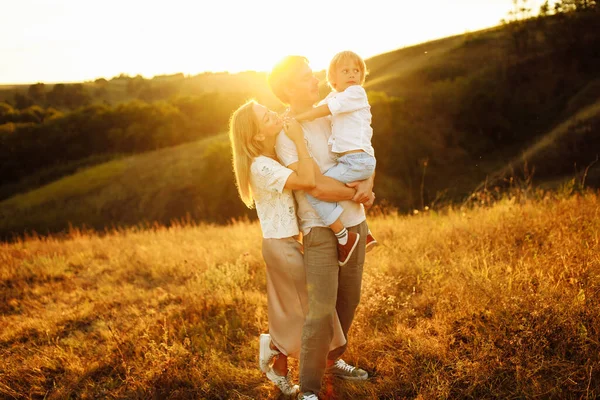 Genitori adorabili con figlio gioioso che cammina sul campo, padre premuroso che tiene il figlio prescolare sulle braccia, madre sorridente e amorevole che abbraccia delicatamente padre bello, genitorialità e concetto di infanzia — Foto Stock