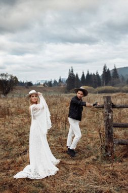 Sahada çitlerin yanında poz veren muhteşem bir çift, şapkalar takan yakışıklı bir adam güzel bir kadına, sevgi dolu bir kocaya bakan güzel bir eş, güzel bir eş, moda fotoshoot konsepti. Yüksek kalite fotoğraf
