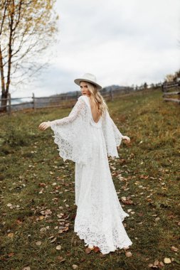 Muhteşem gelin tarlada poz veriyor, sevimli dantel elbise ve beyaz şapka giyiyor, güzel bayan kameraya bakıyor, şefkat ve kadın cazibesi yayıyor, kız güzelliği konsepti