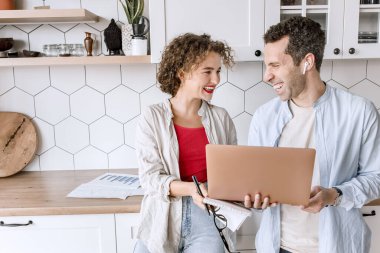 Genç çift evdeki mutfakta dizüstü bilgisayar kullanıyor. 