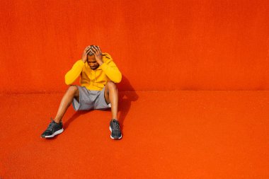 Genç üzgün adam turuncu arka planda yerde oturuyor.