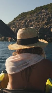 Omuz manzarası, sabit bir kürek çekişini gösteriyor. Çalı çırpı, hasır şapka ve benekli atkı görünür, sıcak arka plan tonları ve mavi su yakınlarındaki sığ odak noktası..