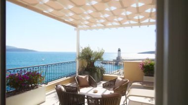 Örülmüş sandalyeleri, küçük bir masası olan güneşli bir sahil terası, turkuaz bir su, dalgalı bir deniz feneri ve parlak gün ışığında uzak bir yelkenliyle karşı karşıya..