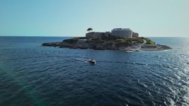 Yuvarlak taştan bir kalesi, geçidi ve rıhtımı olan kayalık bir adanın havadan görünüşü. Derin mavi suların üzerinde parlak, alçak güneş ışığı altında dönen iki motorlu tekne gibi..