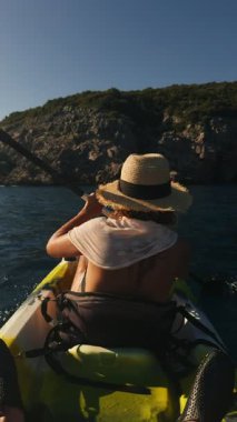 Arka koltuk manzarası, hasır şapkalı bir kürekçinin Karadağ kıyısındaki güneşli kireçtaşı uçurumlarına doğru süzüldüğünü gösteriyor, altın saat ışığı, doymuş blues.