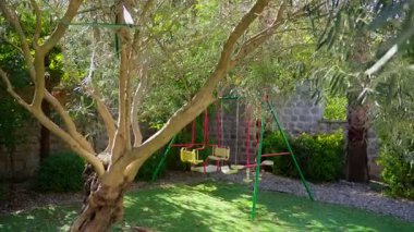 Karadağ 'ın bir bahçesindeki zeytin ağacının altında sarı koltuklu küçük metal bir salıncak bulunuyor ve kamera doğru hareket ediyor. Sabahın köründe benekli ışıklar kabuk ve taşı vurgular..