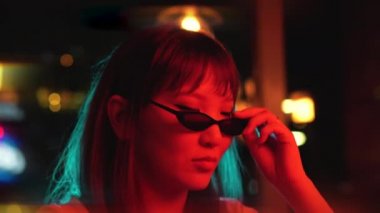 Genç ve güzel bir Asyalı kadının portresi neon ışıkların arka planında. Güneş gözlüklerindeki neon hiyerogliflerin yansıması.