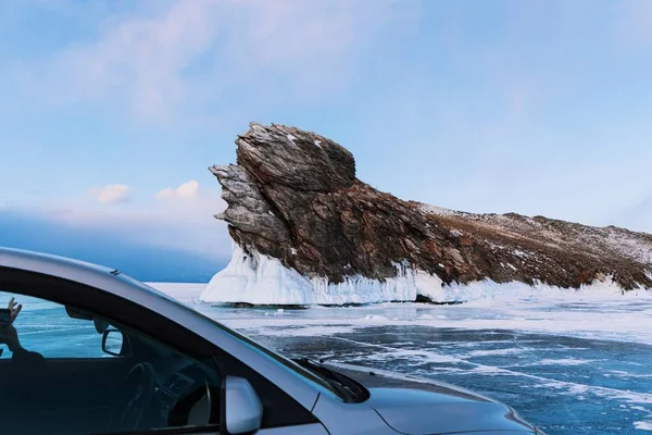 Ogoy Insel auf dem Baikalsee im Winter. Ein graues Auto steht auf glattem Eis vor der Kulisse eines schönen Felsens. Stockbild