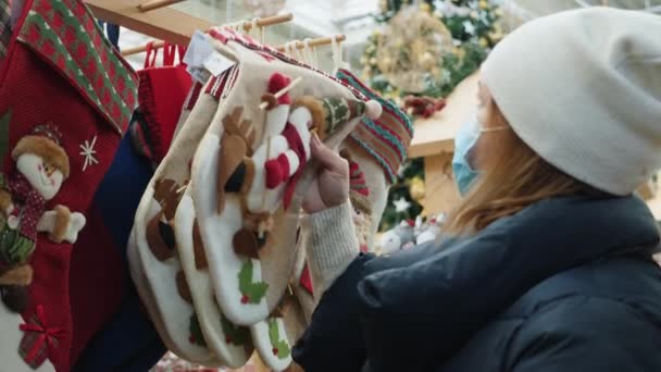 Kvinna i medicinsk mask väljer julstrumpor för gåvor i köpcentret på mässan — Stockvideo