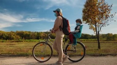 Tıbbi maskeli bir çocukla anne bisikletle yürüyor.